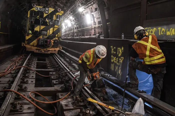 Construction workers in hard hats in the subway tracks, with a jack hammer and shovel, with a work train in the background
